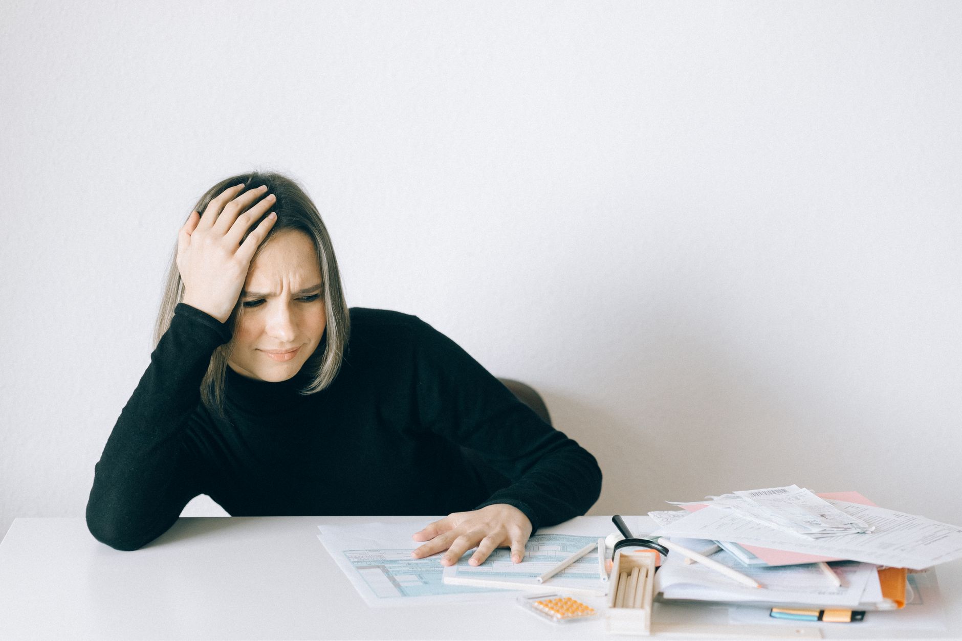 Woman in black sweater stressed with financial paperwork, overwhelmed at white table.