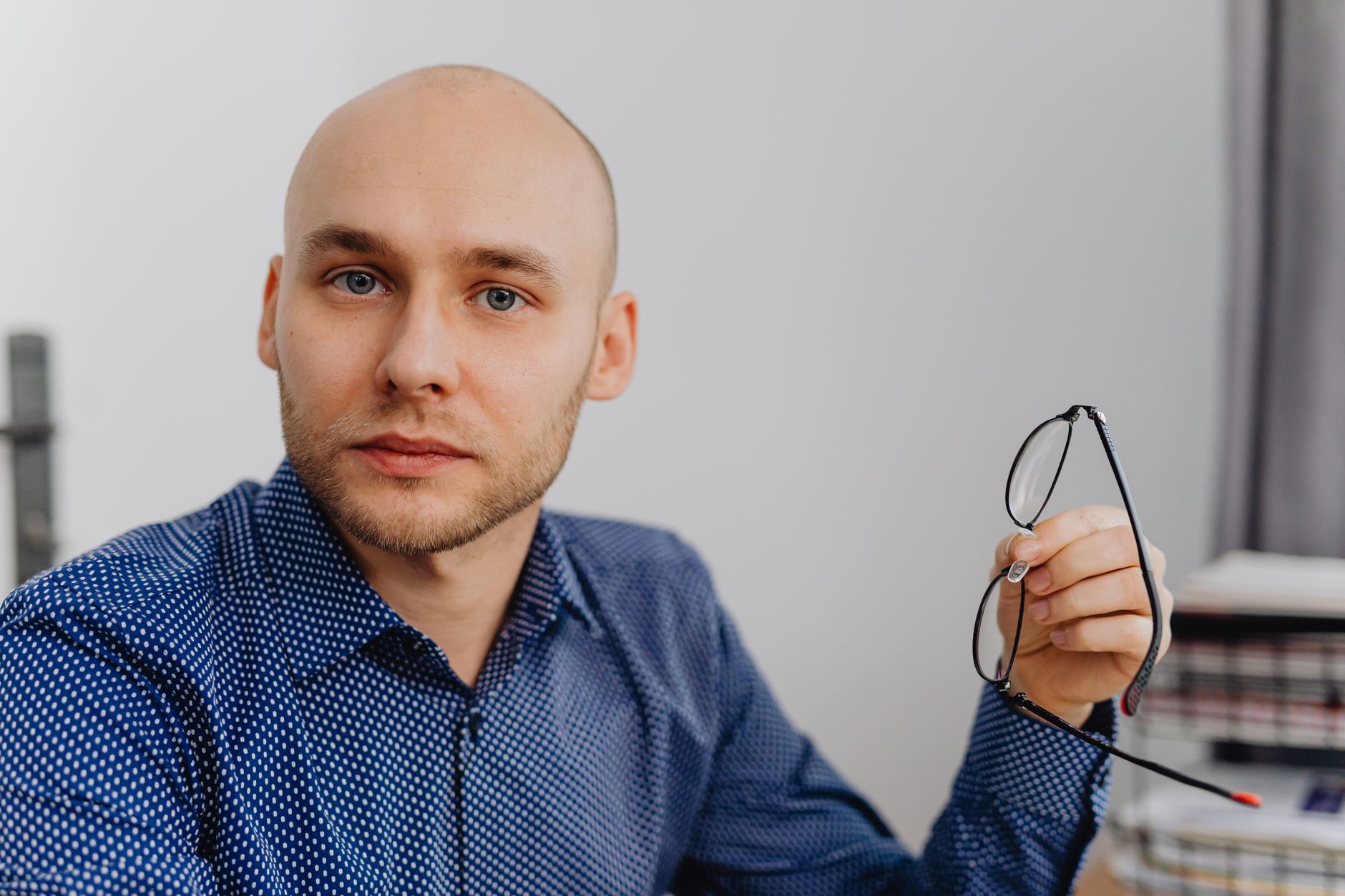 Bald man in a blue shirt holding eyeglasses indoors, with a thoughtful expression.